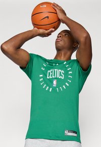 Camiseta de baloncesto verde con texto circular "Celtics" y logo de la NBA. Fabricada con tejido ligero, presenta mangas cortas y un corte recto.