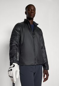 Black puffer jacket with a high collar, zip closure, long sleeves, and a smooth texture. Model holds a white golf glove with black accents.