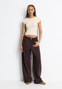 Beige enganierter Kurzarm-Top kombiniert mit lockeren, dunkelbraunen Stoffhosen mit weitem Bein, die einen Kordelzug und einen Knopfverschluss aufweisen.