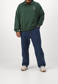 Sweat-shirt vert avec col demi-fermeture éclair, accents de logo brodés. Associé à un jean bleu ample et des baskets blanches avec des semelles beiges.