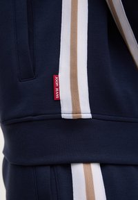 Tissu bleu marine à rayures verticales blanches et beige, avec une étiquette rouge "JOOP! JEANS" cousue près d'une couture de poche.