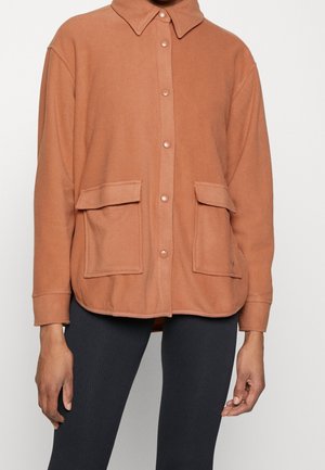Veste marron à boutons avec grandes poches avant, portée sur un pantalon noir, montrée des épaules aux hanches.