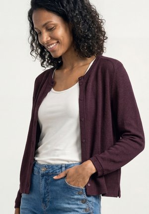 Femme souriante, portant un débardeur blanc, un cardigan violet boutonné et un jean bleu, la main partiellement dans la poche, sur un fond uni.