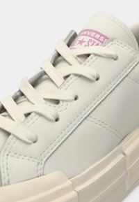 White leather sneaker with cream laces, thick off-white sole, and pink Converse All Star logo on the tongue.