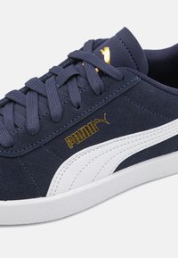 Marineblå semsket sneakers med hvit lærstripe på siden, gulllogo-detaler og flate snørebånd. Strukturerte materialer og lavprofil-design.