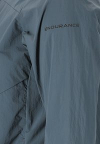 Lätt, strukturerad ljusblå jacka med dragkedjestängning. Har "ENDURANCE"-logotypen i mörkgrått på överarmen.