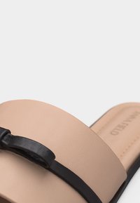 Sandale beige à enfiler avec un nœud et une lanière en cuir noir sur le devant, dotée d'une semelle plate et d'une finition lisse.