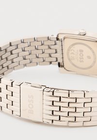 Ασημένιο μεταλλικό μπρασελέ ρολογιού με κούμπωμα χαραγμένο με τη λέξη "BOSS" και οπίσθιο μέρος κάσας ρολογιού της μάρκας "BOSS" μερικώς ορατό.