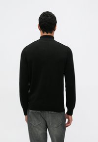 Fekete pulóver bordázott gallérral és mandzsettákkal, sima kötésű textúrával. Szürke farmernadrággal párosítva, hátulról és oldalról látható részletekkel.