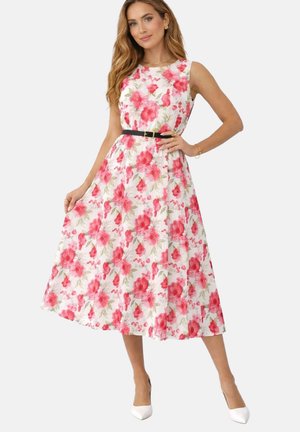 Femme portant une robe midi blanche sans manches avec imprimé floral rouge, ceinture noire et talons blancs, debout avec une main sur la hanche.