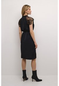 Schwarzes Kleid mit kurzen Spitzenärmel, strukturiertem Stoff, Taillengürtel, knielang und mit seitlichen Schlitzen. Kombiniert mit schwarzen Knöchel-Stiefeln.