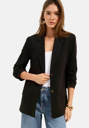 Woman wearing a black blazer over a white top and blue jeans, with rolled-up sleeves and gold bracelet, standing against a plain light background.