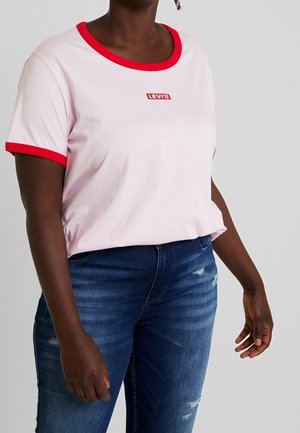 Vrouw draagt een lichtroze Levi's T-shirt met rode kraag en mouwzoom, gecombineerd met blauw versleten spijkerbroek, staand tegen een effen achtergrond.