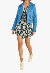 Modrá kožená motorkářská bunda přes zelené šaty s bílými květinovými vzory, doplněná krémovými kotníkovými botami. Hladká textura, dobře padnoucí design.