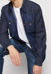 Camisa de denim oscuro con botones a presión, dos bolsillos en el pecho y un yugo de denim más claro en contraste. Llevada sobre una camiseta blanca, con jeans azules desgastados.