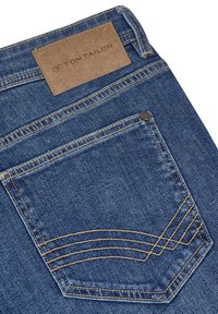 Jeans de denim con un tono azul oscuro, con una etiqueta de cuero marrón que dice "TOM TAILOR". Costura detallada en el bolsillo trasero.