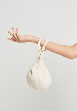 Arm seitlich ausgestreckt mit einer weißen Perlenhandtasche, die am Handgelenk hängt, vor einem einfarbigen hellgrauen Hintergrund.