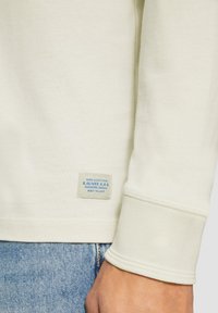 Heller Creme Sweatshirt mit gerippten Bündchen und Saum, versehen mit einem angenähten Etikett, das blaue Schrift zeigt. Das Material hat eine glatte, weiche Textur.