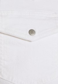 Il tessuto in denim bianco presenta una tasca sul petto con un bottone a pressione metallico argento, accentuata da cuciture tonali e una trama liscia.