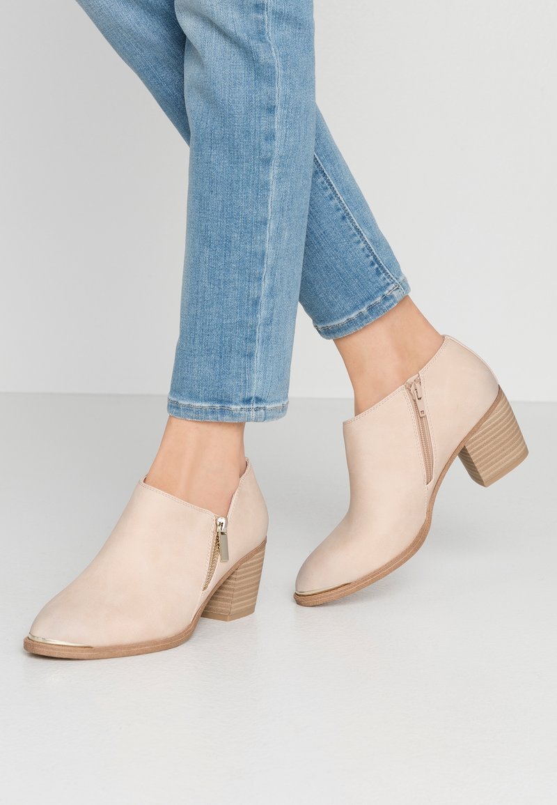 Bottines beige clair à talons carrés avec fermetures éclair latérales, portées avec un jean bleu clair retroussé sur un fond de sol et mur clairs.