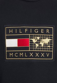 Tessuto nero lavorato a maglia con una toppa rettangolare ricamata che mostra "HILFIGER", una bandiera rossa e bianca, una mappa del mondo dorata e i numeri romani MCMLXXXV.
