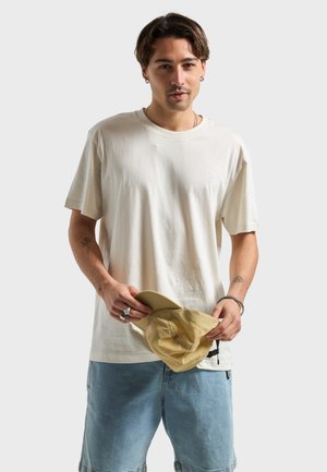 Junger Mann mit Ohrringen und Tattoos hält eine beige Kappe, trägt ein weißes T-Shirt und hellblaue Jeansshorts vor einem einfarbigen Hintergrund.