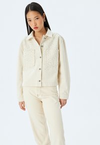 Beige farkkutakki, jossa on niittikoristeet olkapäillä, kaksi etutaskua ja hopeanväriset napit. Yhdistettyyn matching beigenhankkeeseen.