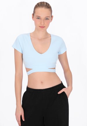 Kobieta o blond włosach w jasnoniebieskim krótkim topie z wycięciami i czarnych spodniach, pozująca z jedną ręką w kieszeni.