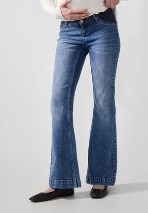 Jeans evazate albastre din denim, cu închidere frontală cu buton, cinci buzunare și o decolorare subtilă pe coapse. Pantofi negri, plata, vizibili.