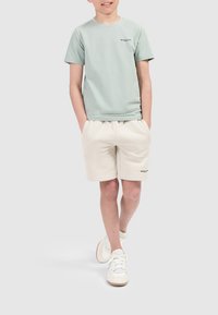 Lichtgroene katoenen t-shirt met korte mouwen en zwart logo; gecombineerd met beige katoenen shorts; witte sneakers met textuurdetails.
