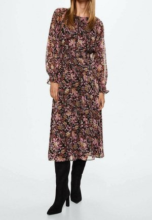 Kobieta ubrana w kwiecistą sukienkę midi z długim rękawem i marszczoną talią, zestawioną z czarnymi wysokimi kozakami na obcasie, na białym tle.