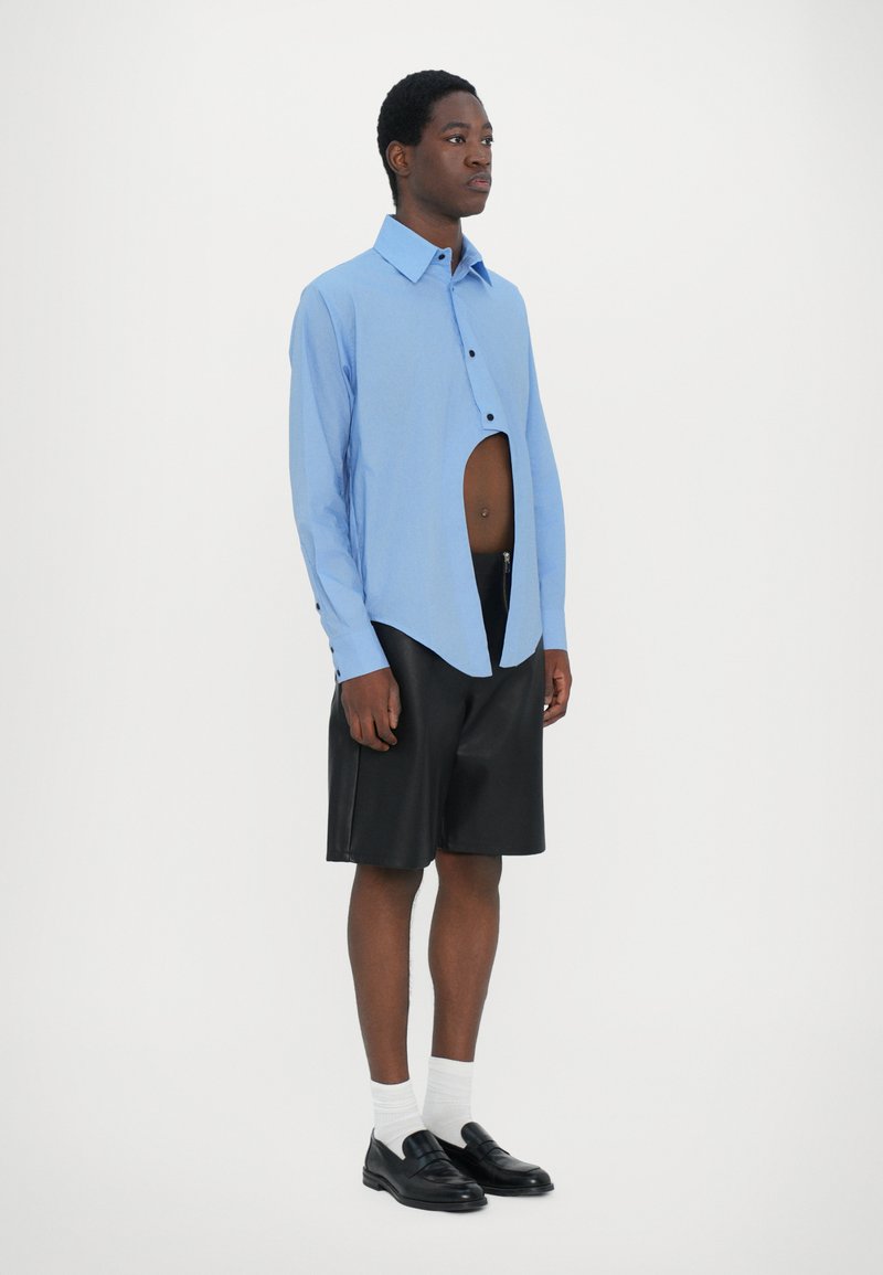 Lyseblå langærmet skjorte med asymmetrisk skæring og knapdetaljer, parret med sorte lædershorts og sorte loafers, hvide strømper.