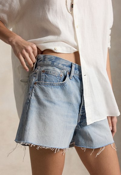 Személy világoskék, kopott farmerrövidnadrágot és fehér, gombos inget visel, az egyik kezét a rövidnadrág derékpántjába süllyesztve.