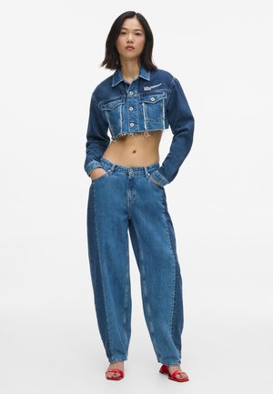 Vrouw die een cropped tweekleurige blauwe spijkerjas en hoge tweekleurige blauwe spijkerbroek draagt, staand met handen in de zakken en rode open teenhakken.