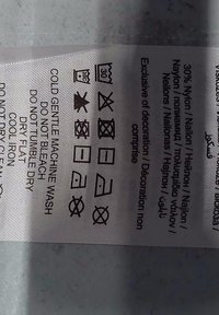 Étiquette d'entretien avec symboles : 30 % nylon. Instructions : lavage en machine, ne pas utiliser d'eau de javel, séchage à plat, repassage à basse température, avec texte multilingue.