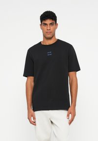 Vīrietis, kurš valkā melnu īso piedurkņu t-kreklu ar nelielu zilu tekstu uz krūtīm un gaišiem biksēm, stāv pretī vienkāršai baltai sienai.