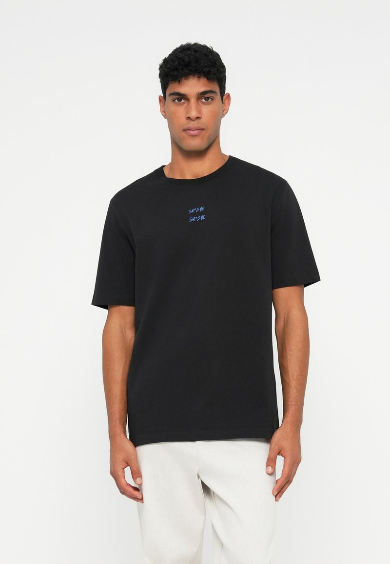 Vīrietis, kurš valkā melnu īso piedurkņu t-kreklu ar nelielu zilu tekstu uz krūtīm un gaišiem biksēm, stāv pretī vienkāršai baltai sienai.