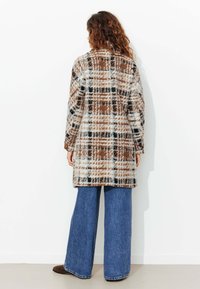Manteau à carreaux en marron, noir et blanc, doté d'un tissu doux et texturé et d'une coupe droite, associé à un pantalon en denim à jambes larges.