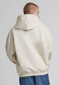 Bé�žová oversized mikina s klokaní kapsou, kapucí na šňůrku a žebrovanými manžetami, v kombinaci s modrými džíny.