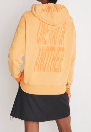 Sudadera naranja con una gran impresión de texto en la parte posterior. Presenta una capucha con cordón y puños de color naranja en contraste. Se lleva con una falda negra.