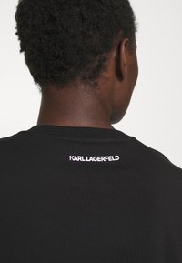 Camiseta negra con cuello redondo, que presenta el texto "KARL LAGERFELD" bordado en rosa en la parte posterior. Textura suave.