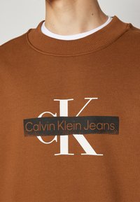 Sweat-shirt marron avec les lettres blanches "cK" et une bande noire traversant la poitrine affichant le texte "Calvin Klein Jeans".
