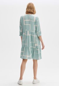 Vaaleanpetroolinsininen mekko, jossa valkoinen abstrakti kuvio, 3/4-hihat, kerätty vyötärö ja kerroksittainen hame, yhdistetty valkoisiin lenkkareihin.