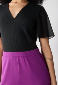 Top nero con scollatura a V e maniche fluttuanti trasparenti, abbinato a pantaloni viola a vita alta. Il tessuto appare liscio con una leggera lucentezza.
