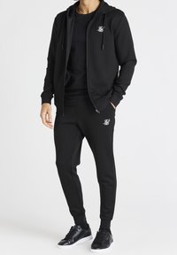 Chándal negro con sudadera con cremallera y pantalones a juego, que presenta un pequeño logo en el pecho y en el tobillo. Tejido suave y liso, con un ajuste entallado.