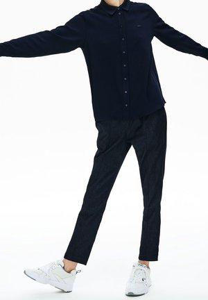 Persona vistiendo una camisa negra abotonada, pantalones negros y zapatillas blancas, de pie con los brazos y las piernas extendidos contra un fondo blanco.