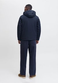 Marineblaues Kapuzenjackett mit strukturiertem Finish, getragen über passenden marineblauen Hosen. Kappenstich-Details und eine lockere Passform. Legere Schuhe.