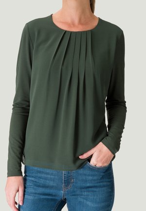 Vrouw draagt een donkergroene blouse met lange mouwen met plooien aan de voorkant en een blauwe spijkerbroek, één hand in de zak, tegen een effen lichte achtergrond.