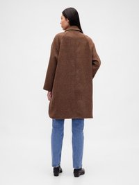 Palton mare, cu carouri maronii, cu guler înclinat, buzunare laterale și o croială lejeră, asortat cu blugi de culoare albastru deschis și încălțăminte neagră.