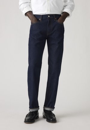 Homme portant un jean bleu foncé retroussé, une chemise blanche boutonnée avec les manches retroussées, des mocassins en cuir noir, les mains dans les poches.